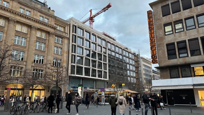 Stadtansicht mit modernem Bau, Menschen auf der Straße und einem Kran im Hintergrund.